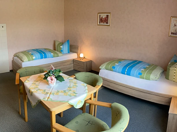 FeWo Schniedermann_Beispielzimmer 2_CC-BY-SA_Teutoburger_Wald_Stadt_Schloss_Holte-Stukenbrock.jpg Zwei Einzelbetten und ein Tisch mit Blumendekor in einem hellen, einfach möblierten Gästezimmer.