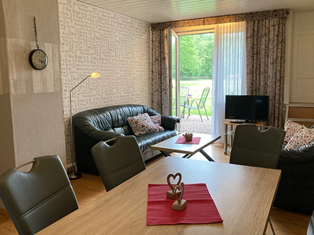 FeWo_Stallmanns Hof_Wohnzimmer_CC-BY-SA_Teutoburger_Wald_Stadt_Schloss_Holte-Stukenbrock.jpg Wohnzimmer mit Ledersofas, offener Terrassentür zum Garten, Fernseher, Holztisch mit Dekoration.