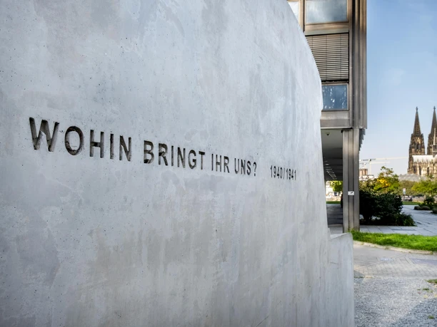 Denkmal der grauen Busse Das Bild zeigt ein Industriedenkmal in Form von "grauen Bussen", eine eindringliche Erinnerung an die tragischen Ereignisse der NS-Zeit. Im Hintergrund ist der Kölner Dom sichtbar. Auf der Betonwand ist die Inschrift "Wohin bringt ihr uns? 1940/1941" zu sehen, die eine tief bewegende Frage stellt.