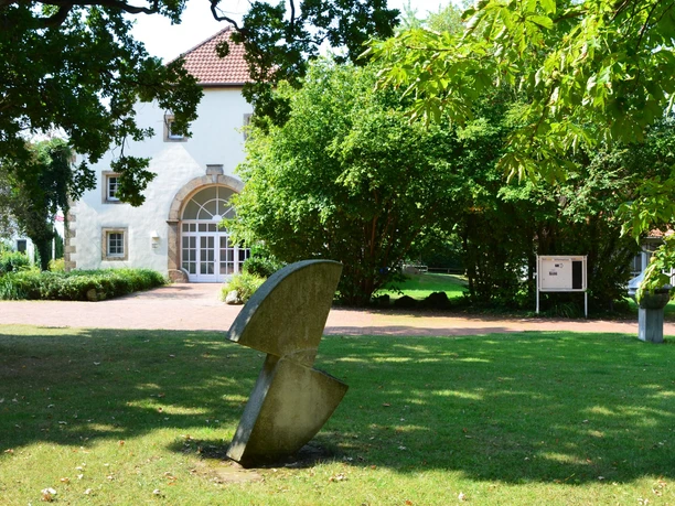 Hotel im Schlosspark_Willebadessen_Aussenaufnahme 1_Avenarius Wohlfühlhotels_2500.jpg Ein modernes Gebäude mit gewölbtem Eingang, umgeben von Laubbäumen und einer abstrakten Skulptur.