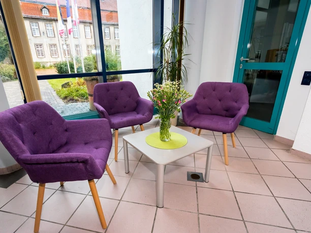Hotel im Schlosspark_Willebadessen_Foyer_Avenarius Wohlfühlhotels.jpg Helles Foyer mit drei lila Sesseln um einen Tisch mit Blumen, Fenster bietet Blick auf Schlosspark.