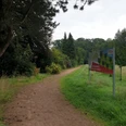 Dreibaechepfad_Babybaumwiese.jpg Ein unbefestigter Weg führt durch einen grünen Park, flankiert von Bäumen und einem Informationsschild.