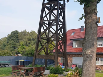 Ein hölzerner Aussichtsturm neben einem Wohnhaus mit rotem Dach, umgeben von Bäumen und Sitzbänken.