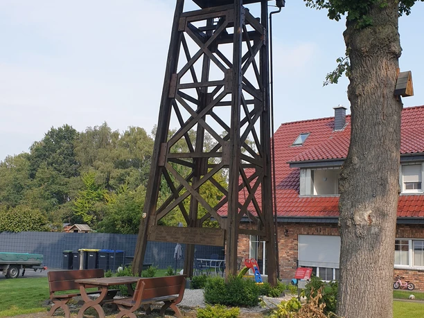 20230830_103956.jpg Ein hölzerner Aussichtsturm neben einem Wohnhaus mit rotem Dach, umgeben von Bäumen und Sitzbänken.