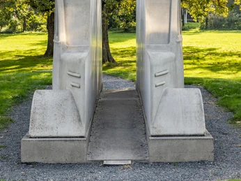 Monument to the gray buses Zwei massive graue Busse aus Beton stehen symbolisch nebeneinander, umgeben von grünem Rasen und Bäumen im Hintergrund.Two massive gray concrete buses stand symbolically next to each other, surrounded by green lawns and trees in the background.