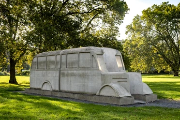 Denkmal der grauen Busse Das Denkmal der grauen Busse zeigt ein stilisiertes, lebensgroßes Betonabbild eines Busses, symbolträchtig in einem Park platziert. Umgeben von Bäumen, fügt sich die Skulptur harmonisch in die grüne Umgebung ein.