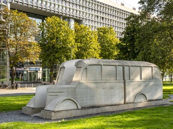 Denkmal der grauen Busse Das Bild zeigt das "Denkmal der grauen Busse" in Köln, eine große steinerne Skulptur in Form eines Busses, umgeben von Bäumen und Rasenflächen im Hintergrund.