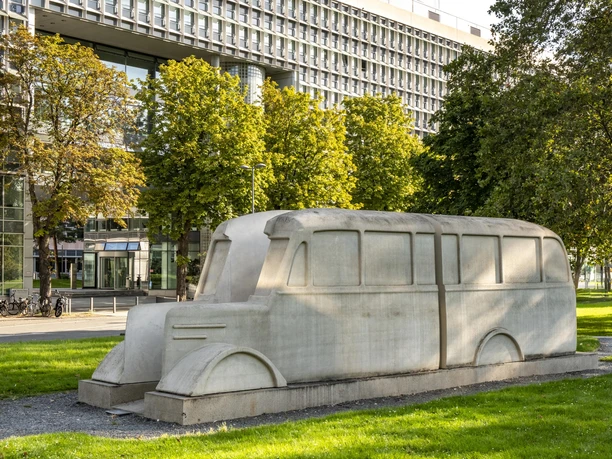 Monument to the gray buses The picture shows the "Monument to the Gray Buses" in Cologne, a large stone sculpture in the shape of a bus, surrounded by trees and lawns in the background.