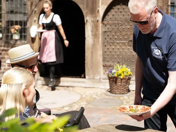 so-schmeckt-bad-zwischenahn-kostprobe-schinken-spieker.jpg Ein Mann serviert einem entspannten Paar im Freien traditionell belegte Ammerländer Schinkenplatte.
