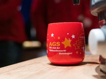 Weihnachtsmarkt Steinhagen AGS Rote Tasse mit Sternenmotiv und goldener Aufschrift "AGS Wir sind der Weihnachtsmarkt Steinhagen!".