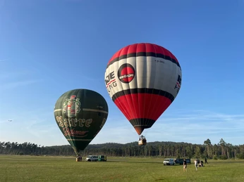 Barre-Broetje-Start-in-Oerlinghausen.jpg Heißluftballons über einer grünen Wiese, Menschen bereiten den Start vor, blauer Himmel im Hintergrund.