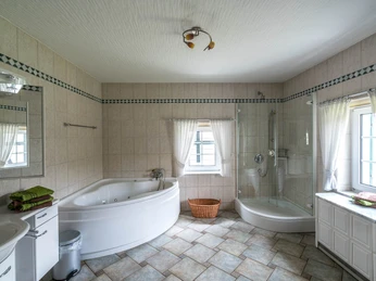 Ennenbach_Badb.jpg Helles Badezimmer mit Eckbadewanne, Dusche, Waschbecken und Fenstern mit weißen Vorhängen.