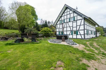 Ennenbach_Garten.jpg Fachwerkhaus in ländlicher Umgebung mit grüner Wiese und altem landwirtschaftlichem Wagen davor.