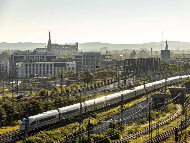 Bahnhof KoelnMesse Deutz Blick auf eine Stadtlandschaft in Köln mit einem Zug, der sich durch die sanft gebogenen Gleise schlängelt, während die ersten Sonnenstrahlen das Panorama erhellen.