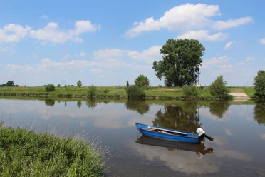 Weser bei Petershagen Windheim Ein ruhiger Flusslauf mit einem blauen Ruderboot im Vordergrund, umgeben von üppigem Grün und einem klaren Himmel mit weißen Wolken.