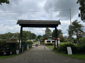 Eingangstor eines Campingplatzes mit Schildern, umgeben von Bäumen und einem wolkigen Himmel.