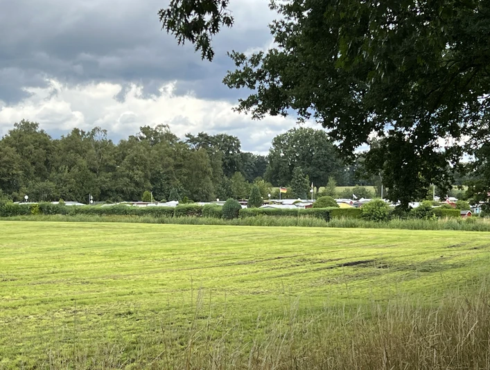 Grüne Wiese mit angrenzendem Campingplatz, umgeben von Bäumen unter einem bewölkten Himmel.