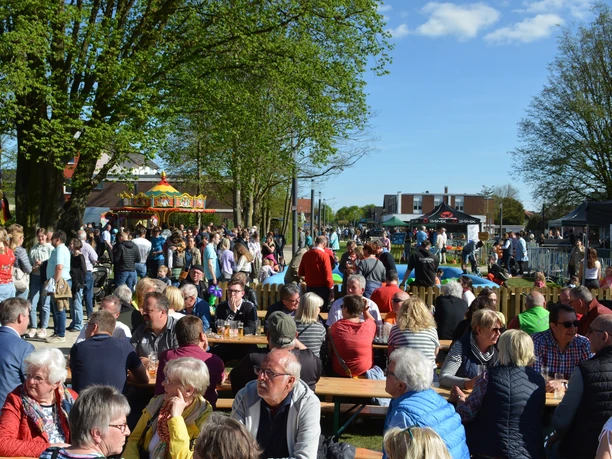 Maibaum- und Radelfest Hövelhof Menschen genießen ein Fest im Freien unter Bäumen, mit Ständen und einem Karussell im Hintergrund.
