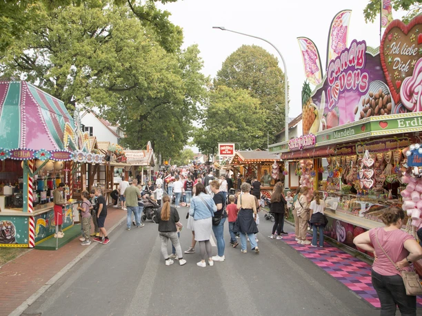 Eine belebte Feststraße, umgeben von bunten Verkaufsständen, voller fröhlicher Marktbesucher.