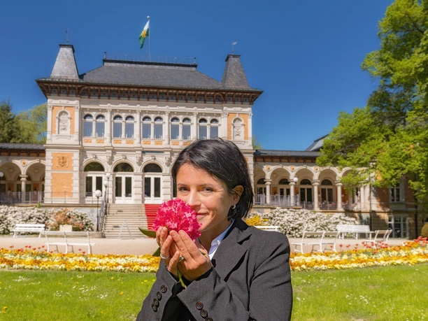 Rhododendronblüte©JanBräuer.jpg Eine Frau hält eine Rhododendronblüte vor einem historischen Gebäude.