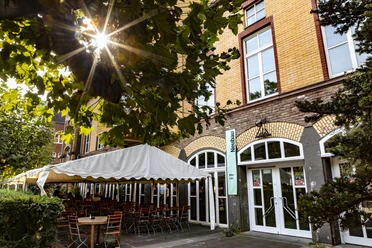 Alte Feuerwache The Alte Feuerwache has a historic façade made of yellow bricks with a high window front. A parasol shades numerous outdoor seats, framed by green plants.