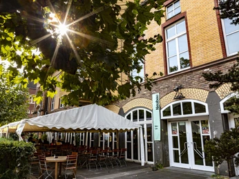 Alte Feuerwache Die Alte Feuerwache hat eine historische Fassade aus gelben Ziegeln mit hoher Fensterfront. Ein Sonnenschirm beschattet zahlreiche Außenplätze, eingerahmt von grünen Pflanzen.The Alte Feuerwache has a historic façade made of yellow bricks with a high window front. A parasol shades numerous outdoor seats, framed by green plants.