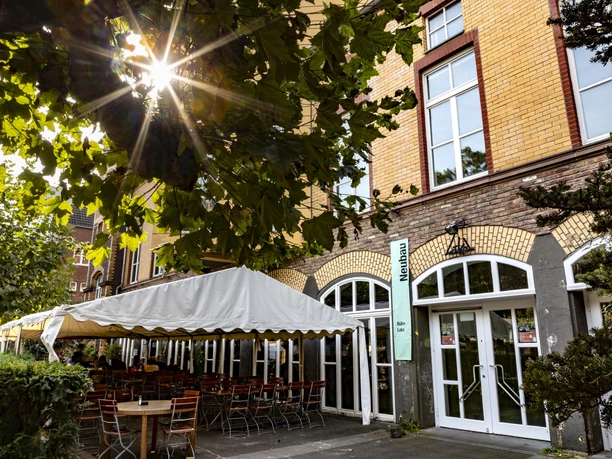 Alte Feuerwache Die Alte Feuerwache hat eine historische Fassade aus gelben Ziegeln mit hoher Fensterfront. Ein Sonnenschirm beschattet zahlreiche Außenplätze, eingerahmt von grünen Pflanzen.
