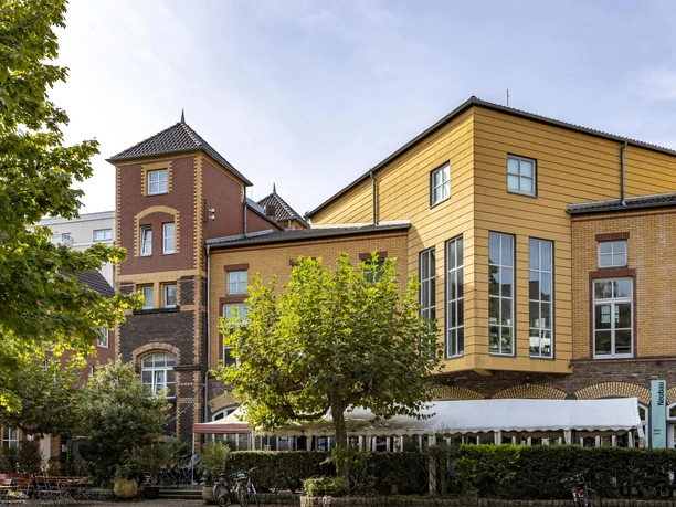 Alte Feuerwache Alte Feuerwache Köln mit einer markanten Kombination aus Backsteinfassaden und modernen Holzelementen in warmen Gelbtönen. Im Vordergrund stehen Bäume, die dem Gebäude eine grüne Umrahmung verleihen.