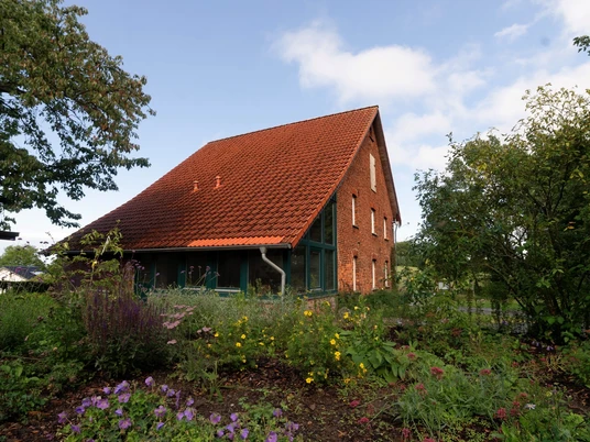 Ein rotes Backsteinhaus mit Satteldach, umgeben von blühenden Pflanzen und Bäumen unter blauem Himmel.