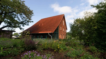 lippischeauszeit.jpg Ein rotes Backsteinhaus mit Satteldach, umgeben von blühenden Pflanzen und Bäumen unter blauem Himmel.
