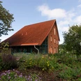 lippischeauszeit.jpg Ein rotes Backsteinhaus mit Satteldach, umgeben von blühenden Pflanzen und Bäumen unter blauem Himmel.