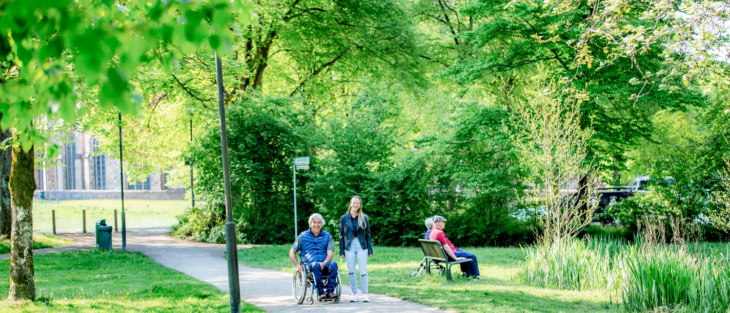 Spaziergang in Altenberg <p>Ein Rollstuhlfahrer wird von einer Frau auf einem sonnigen Parkweg begleitet, während eine andere Person auf einer Bank sitzt.</p>