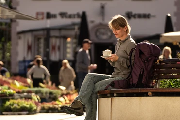 Innenstadt Wipperfürth <p>Frau genießt Kaffee auf einer Bank bei sonnigem Wetter in belebter Straßenatmosphäre.</p>