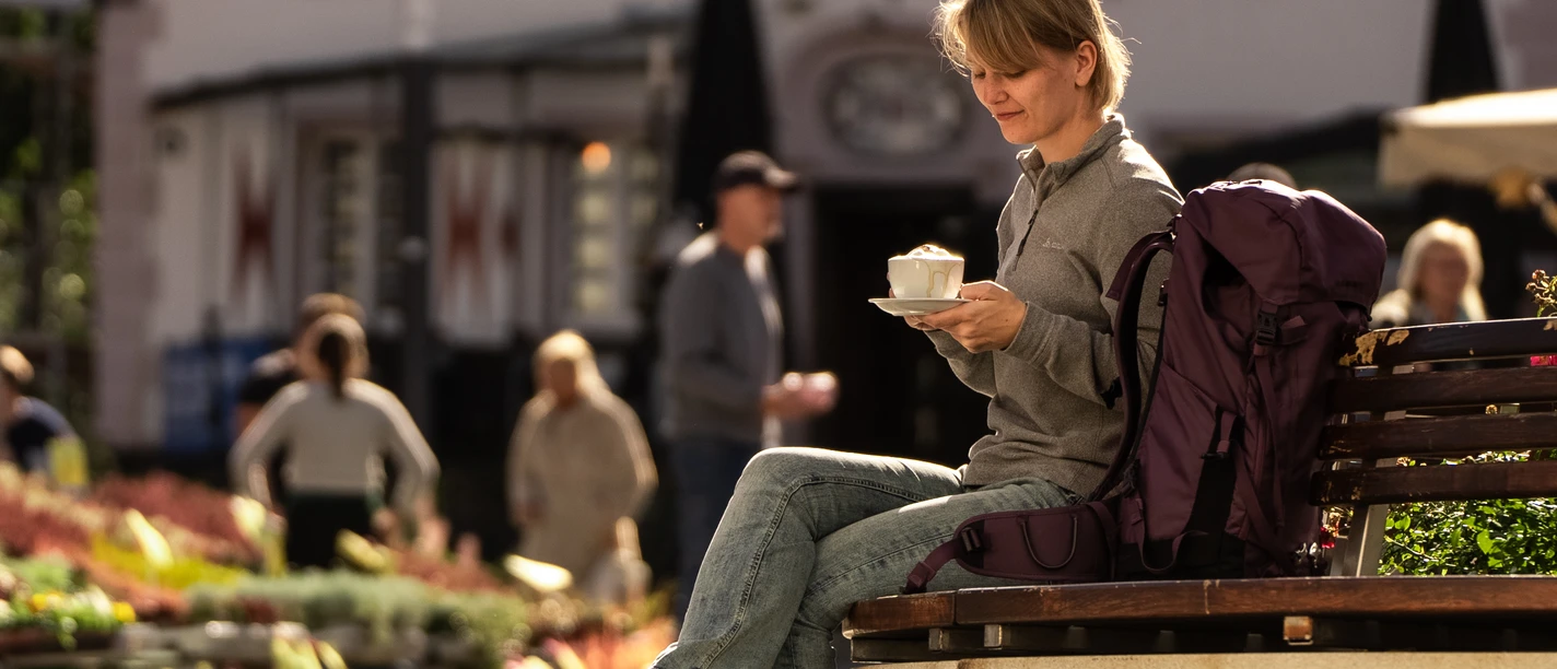 Innenstadt Wipperfürth <p>Frau genießt Kaffee auf einer Bank bei sonnigem Wetter in belebter Straßenatmosphäre.</p>