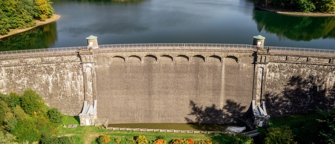 Neyetalsperre Staudamm in idyllischer Landschaft, umgeben von Wald und Wasser, unter blauem Himmel.