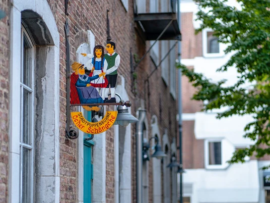 Buntes Puppentheater-Schild „Öcher Schängche“ an einer Backsteinfassade. Zwei stilisierte Puppen sind darauf abgebildet.