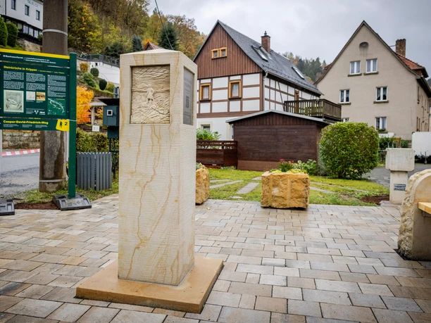 Stone column with relief on paved square, surrounded by half-timbered houses and information board; autumnal atmosphere.