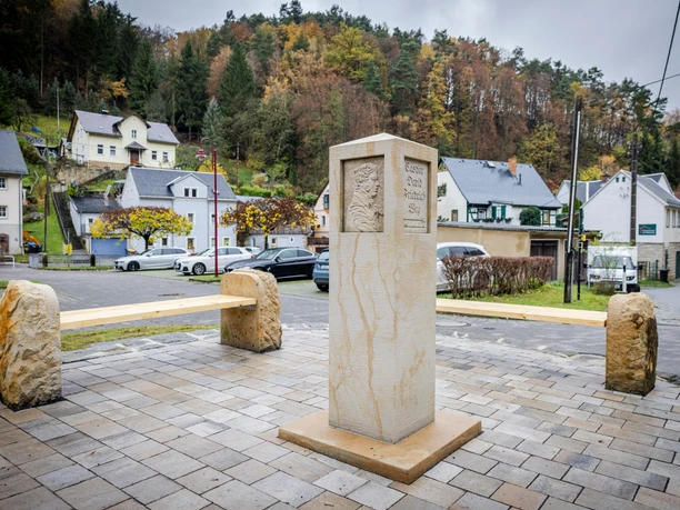 Stone bench and monument on a paved square, surrounded by cars and houses, with a wooded hill in the background on a cloudy day.