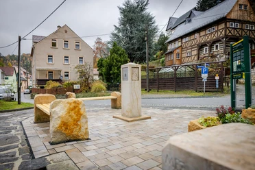 Stenen bank en monument op een geplaveid plein, omringd door een vakwerkhuis en woongebouw, onder een bewolkte hemel.