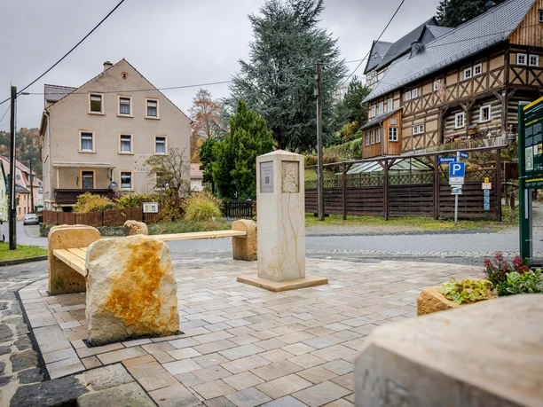Stone bench and monument on a paved square, surrounded by a half-timbered house and residential building, under a cloudy sky.