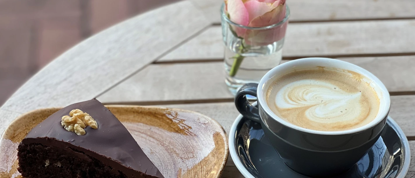 A slice of chocolate cake with walnuts and a latte macchiato next to a vase of roses on wood.