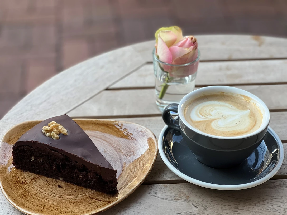 Kaffee und Kuchen (1) Ein Stück Schokoladenkuchen mit Walnuss und ein Latte Macchiato neben einer Rosenvase auf Holz.