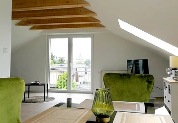 Helles Wohnzimmer mit großen Fenstern, grünen Sesseln, Tischplatte und Dachschräge mit Holzbalken.