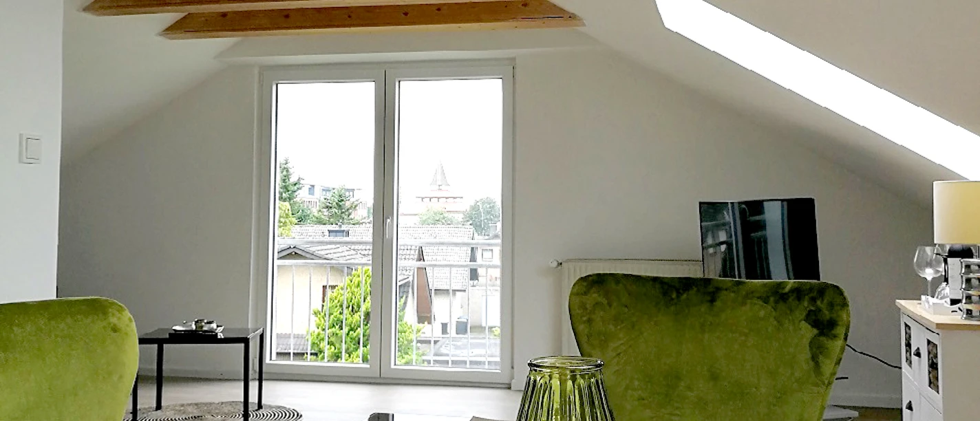 Helles Wohnzimmer mit großen Fenstern, grünen Sesseln, Tischplatte und Dachschräge mit Holzbalken.