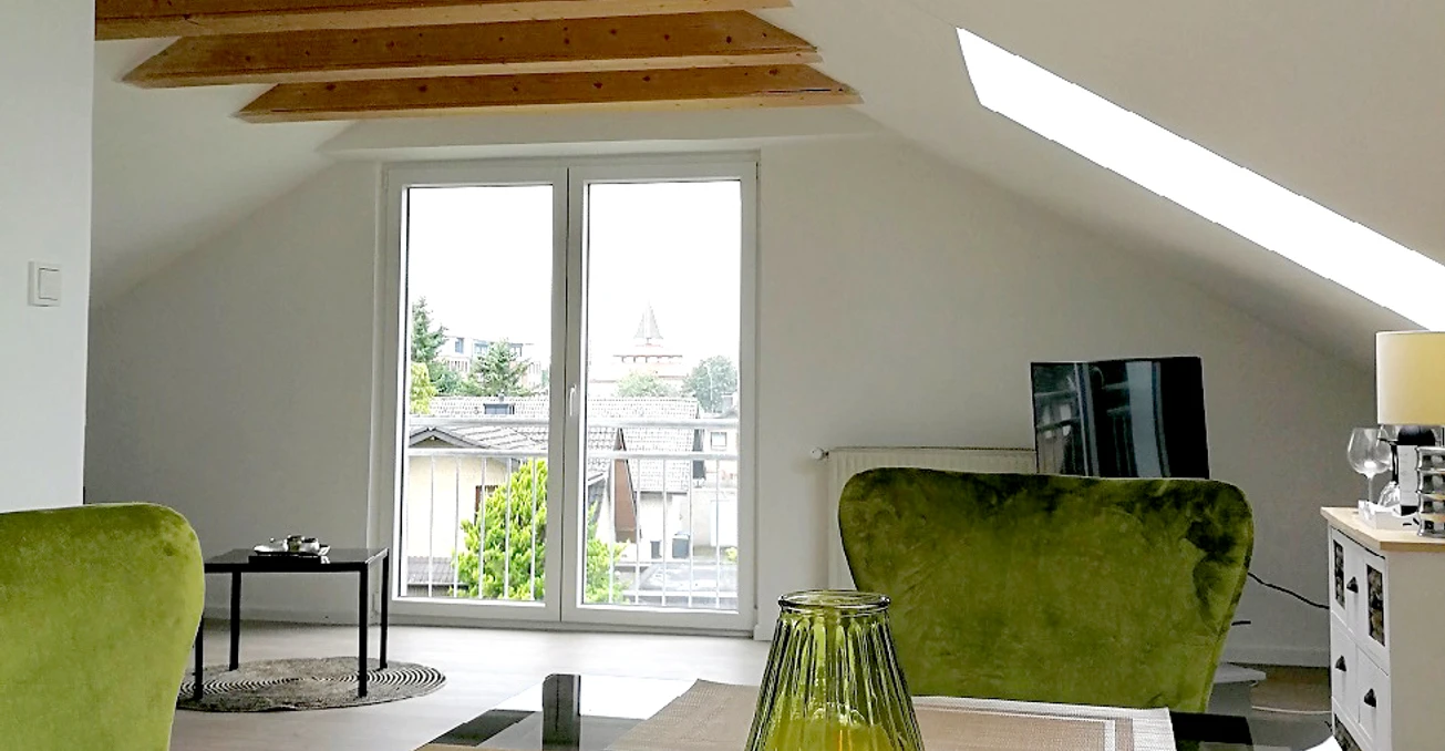 FW_Berliner Straße _b© U_Kleinehagenbrock.jpg Helles Wohnzimmer mit großen Fenstern, grünen Sesseln, Tischplatte und Dachschräge mit Holzbalken.