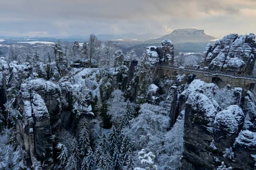 Basteibrücke Verschneite Felsformationen und eine steinerne Brücke in einer winterlichen Landschaft, umgeben von schneebedeckten Bäumen und einem bewölkten Himmel.