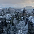 Basteibrücke Verschneite Felsformationen und eine steinerne Brücke in einer winterlichen Landschaft, umgeben von schneebedeckten Bäumen und einem bewölkten Himmel.Snow-covered rock formations and a stone bridge in a wintry landscape, surrounded by snow-covered trees and a cloudy sky.Zasněžené skalní útvary a kamenný most v zimní krajině, obklopené zasněženými stromy a zataženou oblohou.Pokryte śniegiem formacje skalne i kamienny most w zimowym krajobrazie, otoczone ośnieżonymi drzewami i zachmurzonym niebem.Besneeuwde rotsformaties en een stenen brug in een winters landschap, omringd door besneeuwde bomen en een bewolkte lucht.Formazioni rocciose innevate e un ponte di pietra in un paesaggio invernale, circondato da alberi innevati e da un cielo nuvoloso.