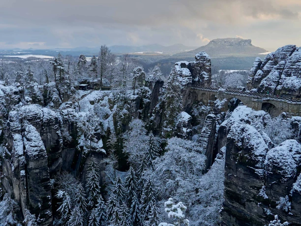 Basteibrücke Verschneite Felsformationen und eine steinerne Brücke in einer winterlichen Landschaft, umgeben von schneebedeckten Bäumen und einem bewölkten Himmel.