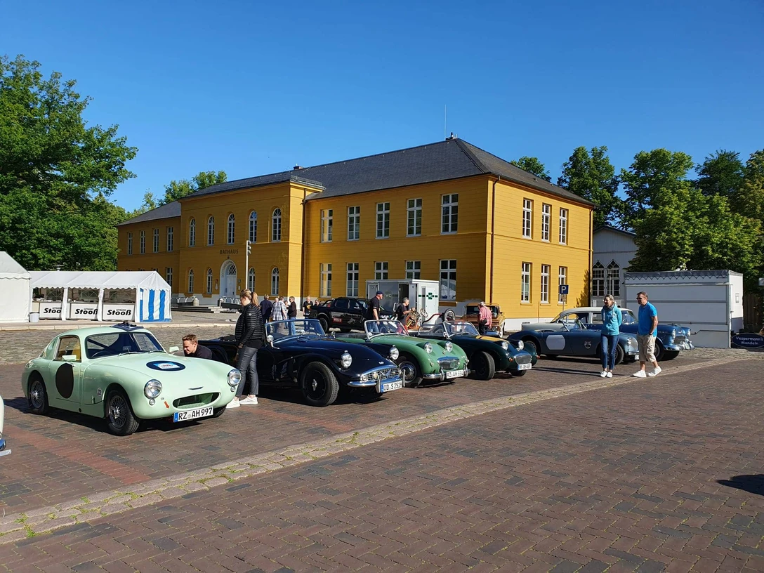 Rallye_Oldtimer.jpg Mehrere Oldtimer stehen vor dem Rathaus auf dem Parkplatz. Menschen schauen sie interessiert an