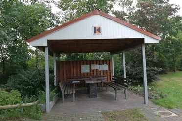 Kolping Schutzhütte 3.jpg Überdachte Raststelle mit Holzbänken und Tisch, umgeben von Bäumen und Grünflächen in Bawinkel.